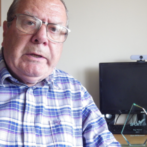 A man in a checkered shirt sits in front of a desk with his Sported volunteer award behind him