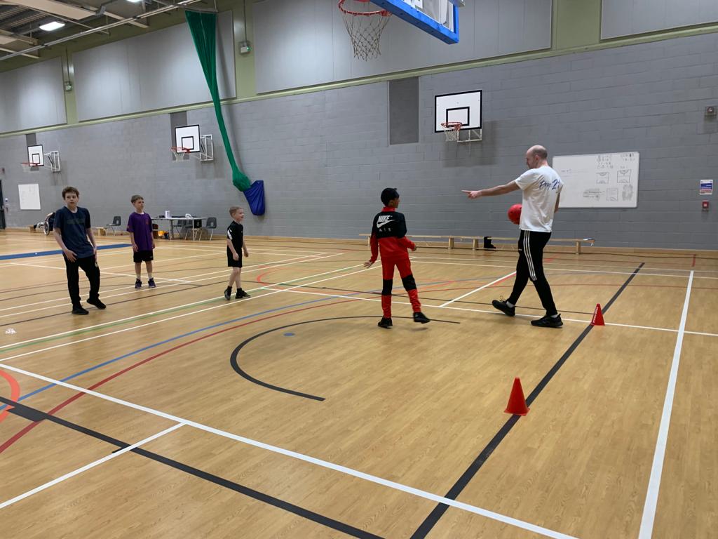 A group of young boys are in a sports hall being instructed by a man holding a ball.