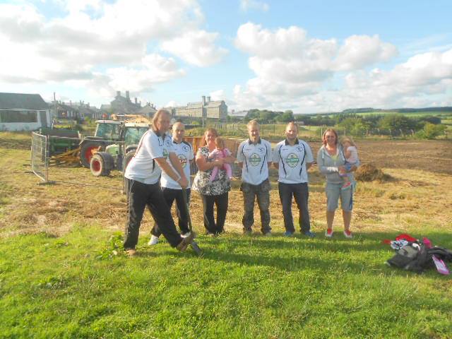 Six adults, some wearing football shirts, two women holding babies. Standing in a field with a digger and other plant equipment. One man is posing with a spade digging into the grass