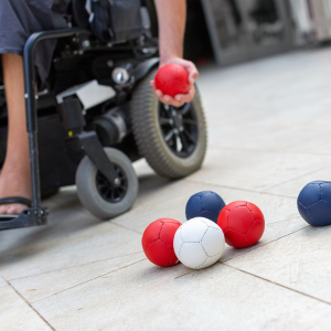 close up of a power wheelchair with person holding a boccia ball. Red, white and blue boccia balls in foreground