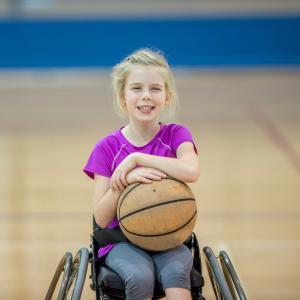 smiling girl holding a basket ball in a wheelchair, sports court in the background